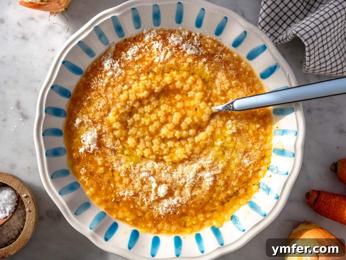 Soothing Small Pasta 4 A close-up of creamy Pastina soup with grated Parmesan cheese, served with a blue spoon in a white bowl.