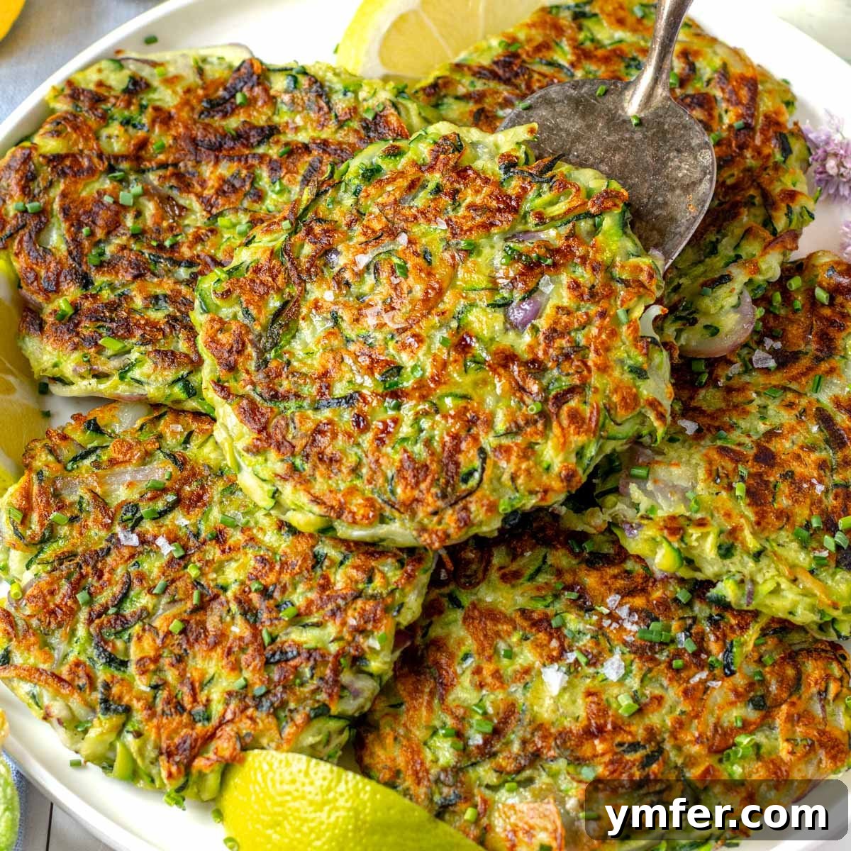 Golden brown zucchini fritters artfully arranged on a white plate with fresh lemon wedges.