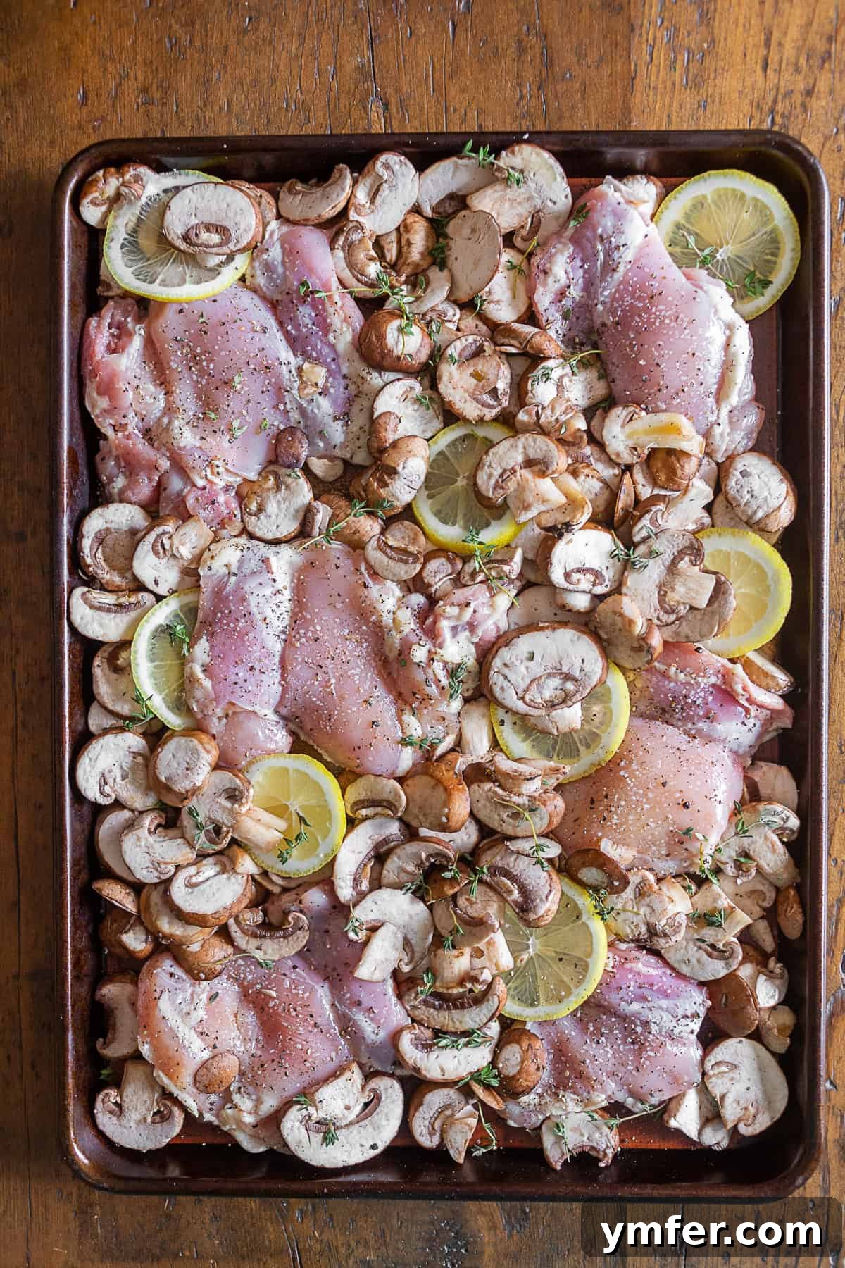 One Pan Zesty Lemon Garlic Chicken 4 Uncooked ingredients for Sheet Pan Garlic Lemon Chicken and Mushrooms arranged on a baking sheet: raw chicken thighs, sliced lemons, baby portobello mushrooms, and fresh thyme sprigs.