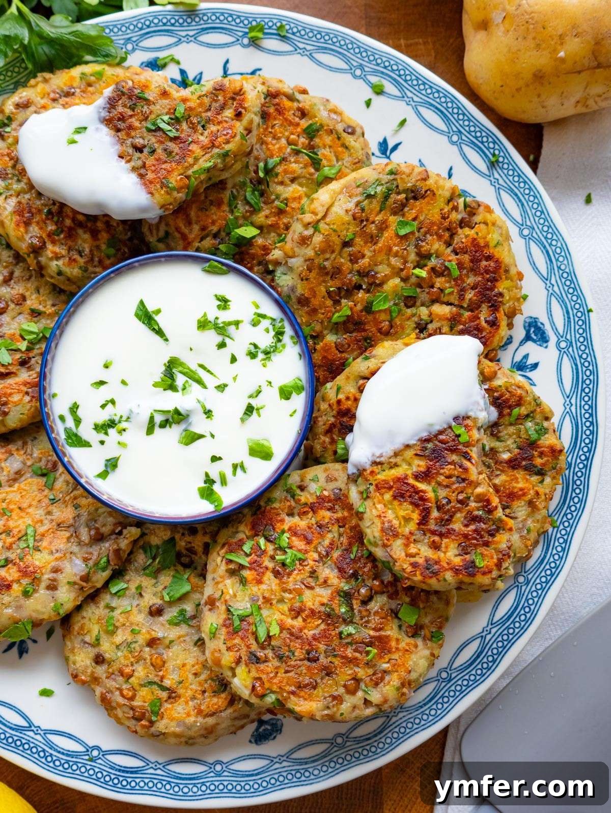 A top-down view of freshly cooked golden-brown lentil potato patties, neatly arranged on a white plate around a small bowl of creamy yogurt dip, garnished with fresh herbs.