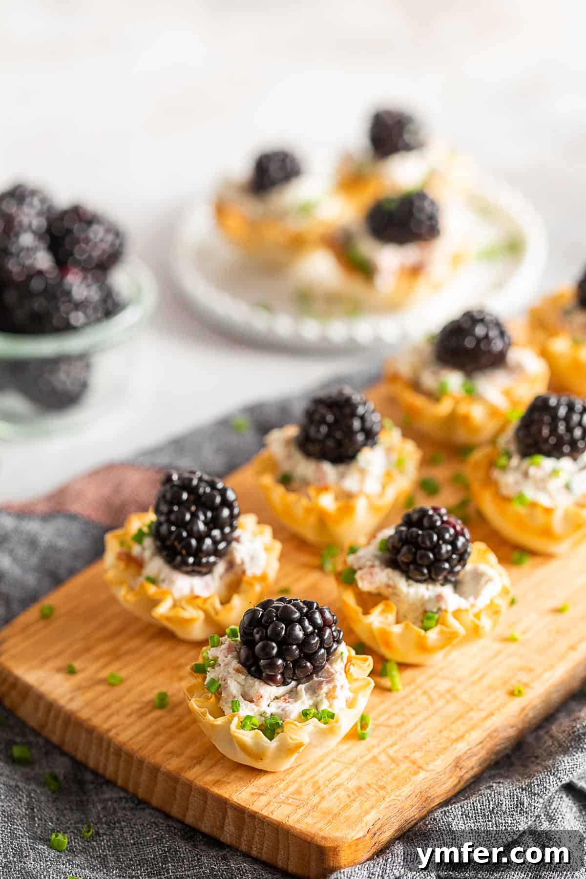 Golden Phyllo Goat Cheese Prosciutto Parcels 2 Goat Cheese Prosciutto Phyllo Bites arranged beautifully on a rustic wooden board, garnished with fresh chives and blackberries.