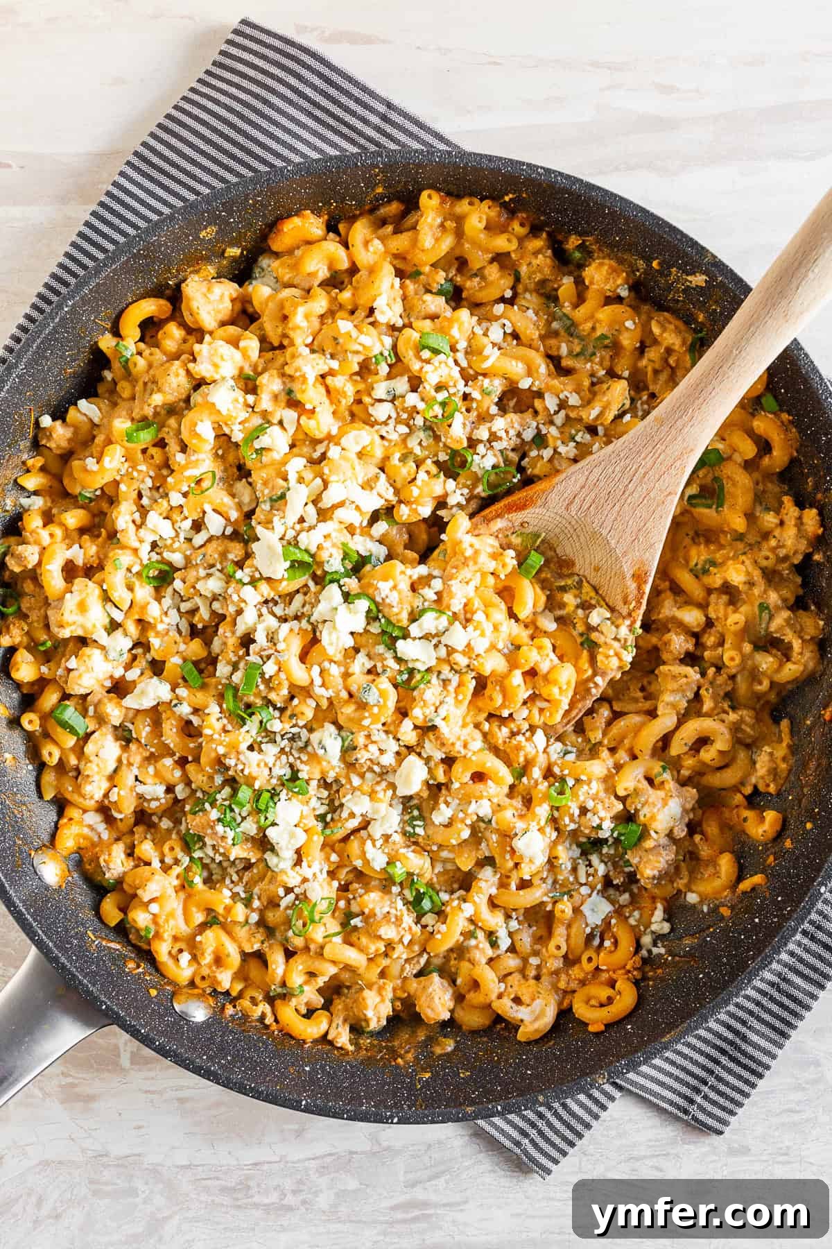 Creamy One Pot Buffalo Chicken Pasta 2 A large skillet filled with creamy One Pot Buffalo Chicken Pasta, garnished with fresh blue cheese crumbles and chopped green onions. A wooden spoon rests in the skillet, inviting a taste.