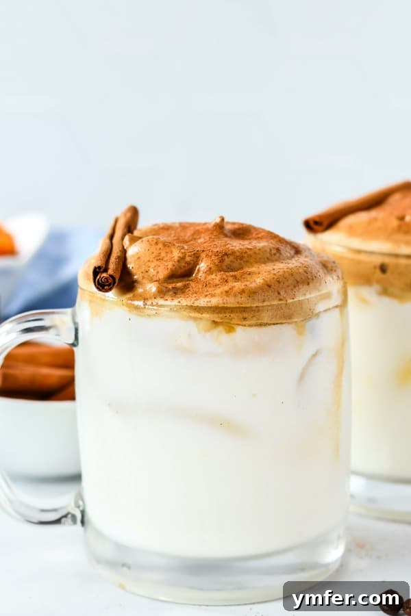 A glass mug filled with frothy pumpkin spice whipped coffee, topped with a sprinkle of cinnamon, sitting on a wooden table with fall decorations.