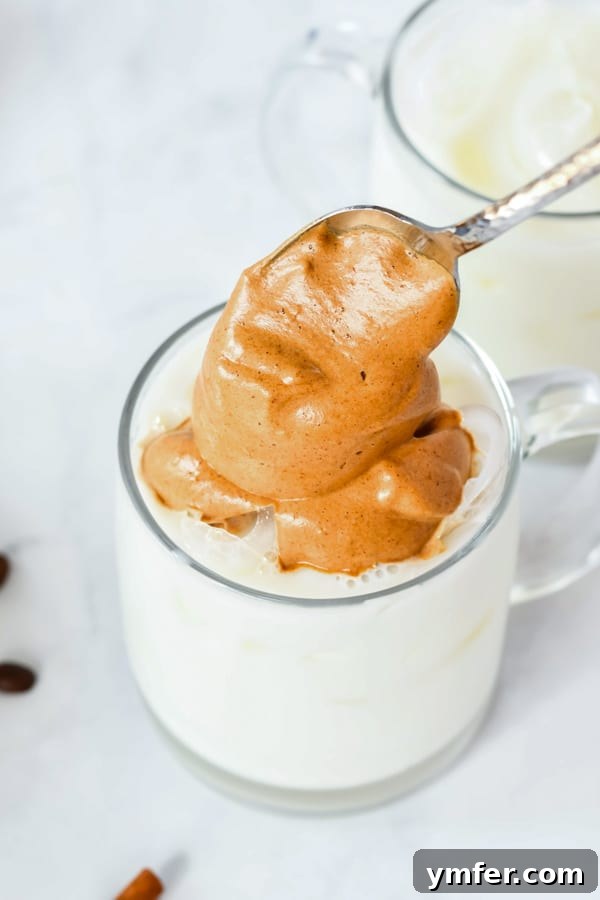 A hand gently adding the creamy pumpkin spice whipped coffee foam to a glass mug filled with ice and milk, showcasing the beautiful layered effect.