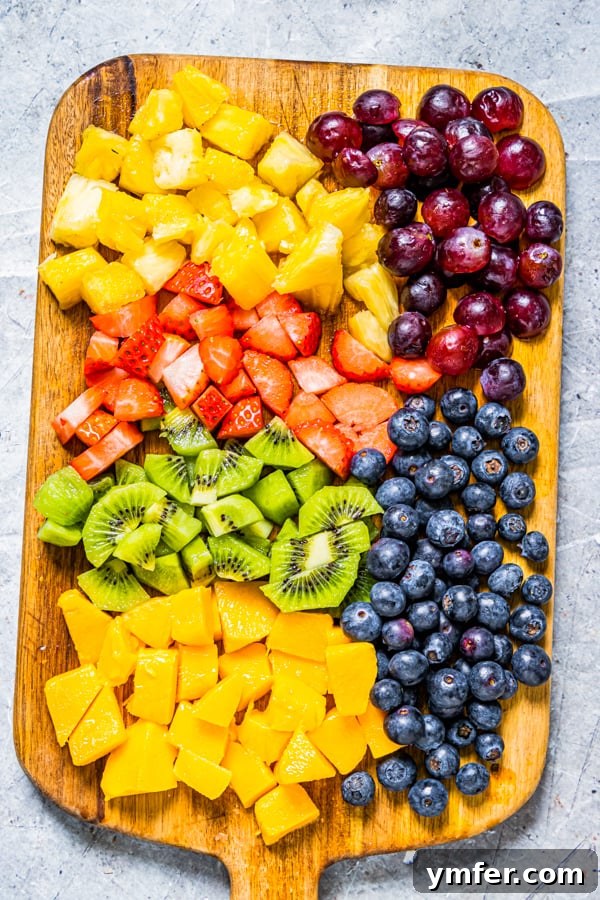 A vibrant assortment of freshly chopped fruits including strawberries, mango, pineapple, kiwi, blueberries, and red grapes neatly arranged on a wooden cutting board, ready for popsicle making.