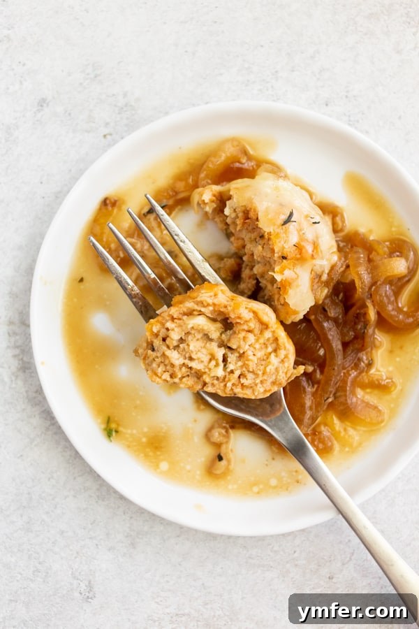 A French Onion Chicken Meatball cut in half on a plate with a fork, showing its tender interior and rich sauce. An inviting close-up.