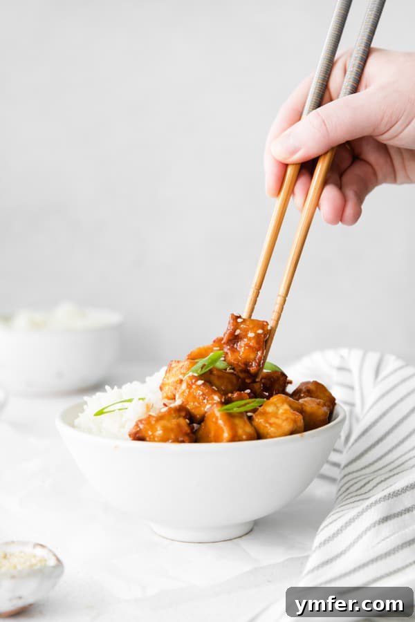Crispy Sweet Chili Air Fryer Tofu 3 A person holding chopsticks, picking up a piece of crispy Air Fryer Sweet Chili Tofu from a bowl.
