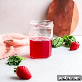 Fresh Strawberry Basil Elixir 10 Small bottle of homemade strawberry basil simple syrup, garnished with fresh strawberries and basil leaves, on a light background.