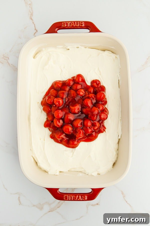 Luscious Cherry Cheesecake Dip 4 A smooth layer of cherry pie filling being evenly spread over the prepared creamy cheesecake dip in a baking dish, creating a vibrant, appealing topping.