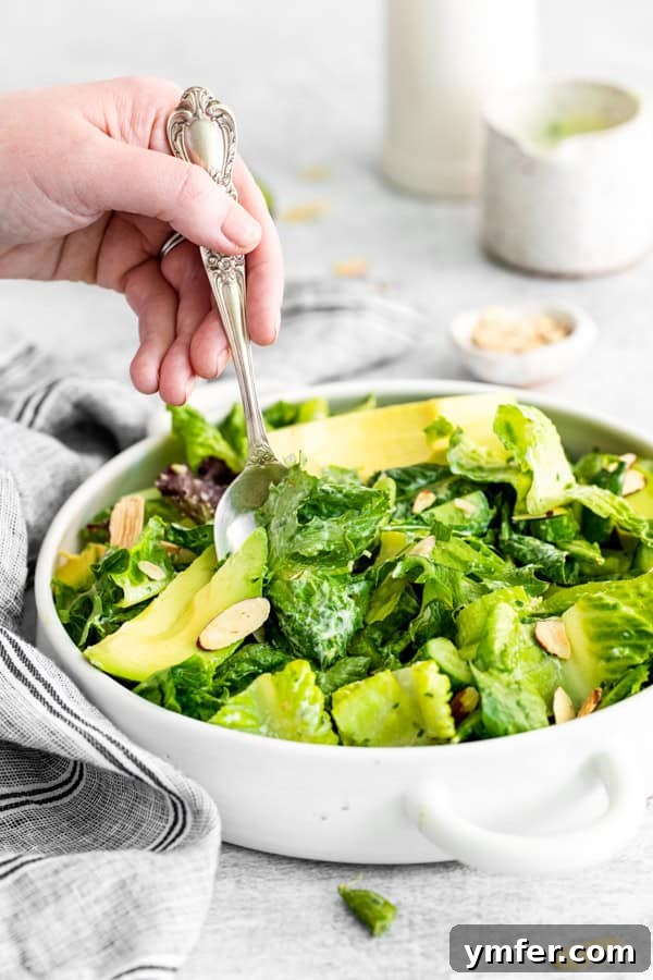Divine Green Goddess Salad 4 A close-up of a hand serving a spoonful of Green Goddess Salad from a large bowl, highlighting the vibrant greens, avocado, and dressing.