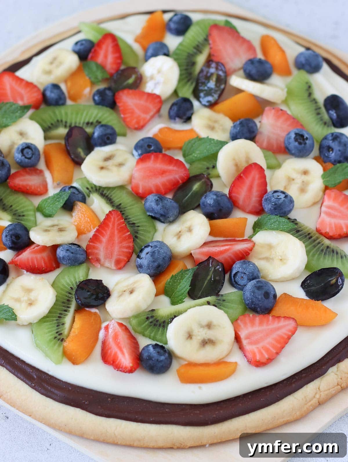 Rainbow Fruit Pizza 13 A close-up of a fruit pizza, showcasing a delightful topping mix of fresh strawberries, grapes, blueberries, banana slices, and kiwi.
