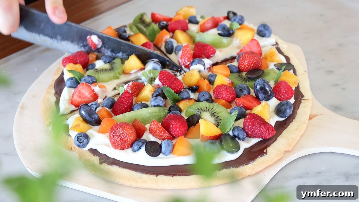 Rainbow Fruit Pizza 3 A hand gently slicing into a vibrant fruit pizza, revealing its crunchy sugar cookie base, creamy dairy-free frosting, and colorful fresh fruit topping.