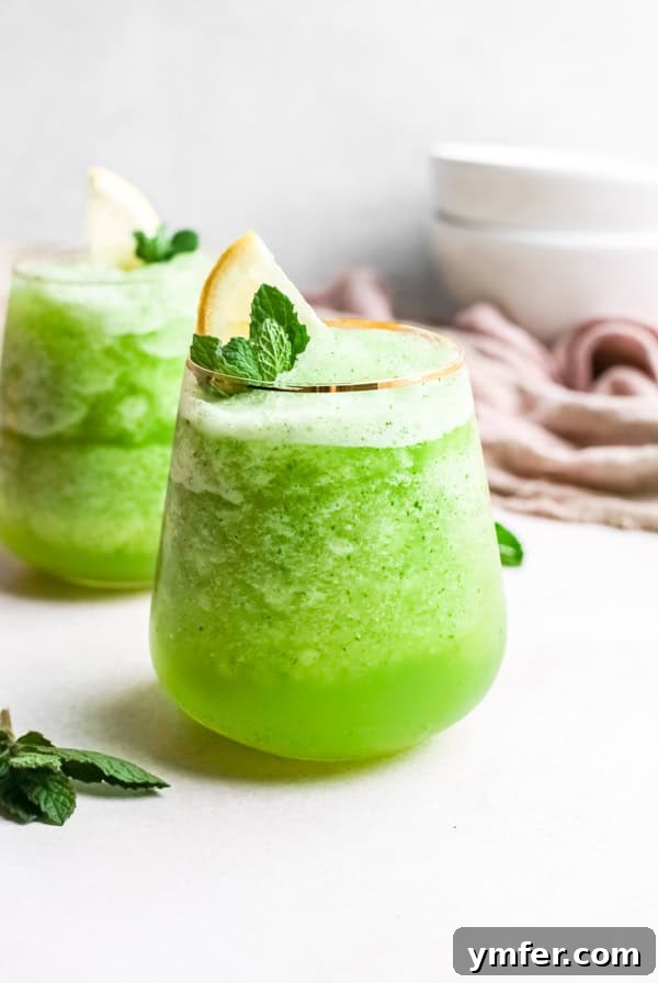 Limonana Frost 2 A tall glass of homemade frozen mint lemonade (Limonana) garnished with fresh mint leaves and a lemon slice, sitting on a light surface with a blurred background.