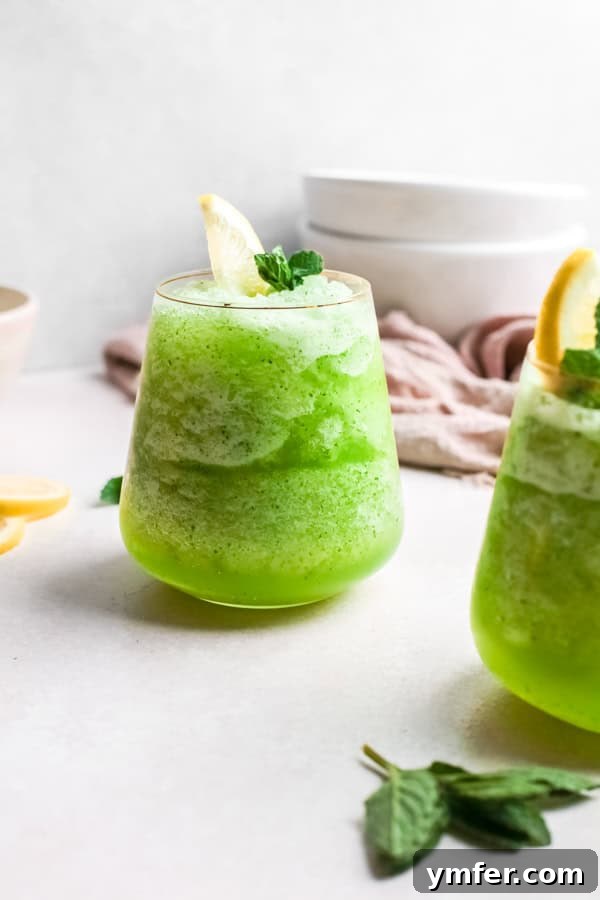 Limonana Frost 5 Two glasses of frozen mint lemonade (Limonana) with garnishes of mint and lemon, served on a light table.