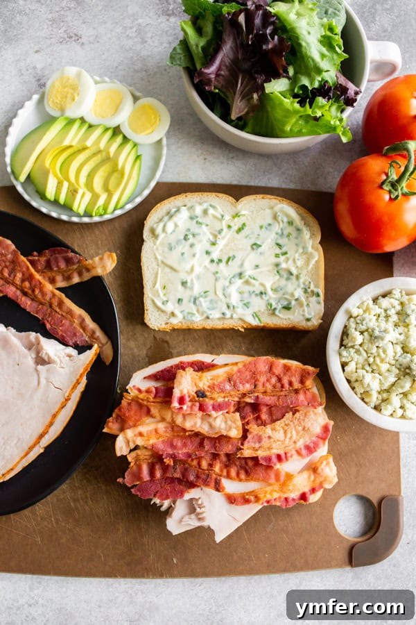 The Ultimate Cobb Sandwich 4 A Cobb Salad Sandwich being assembled on a wooden cutting board, with ingredients carefully layered.