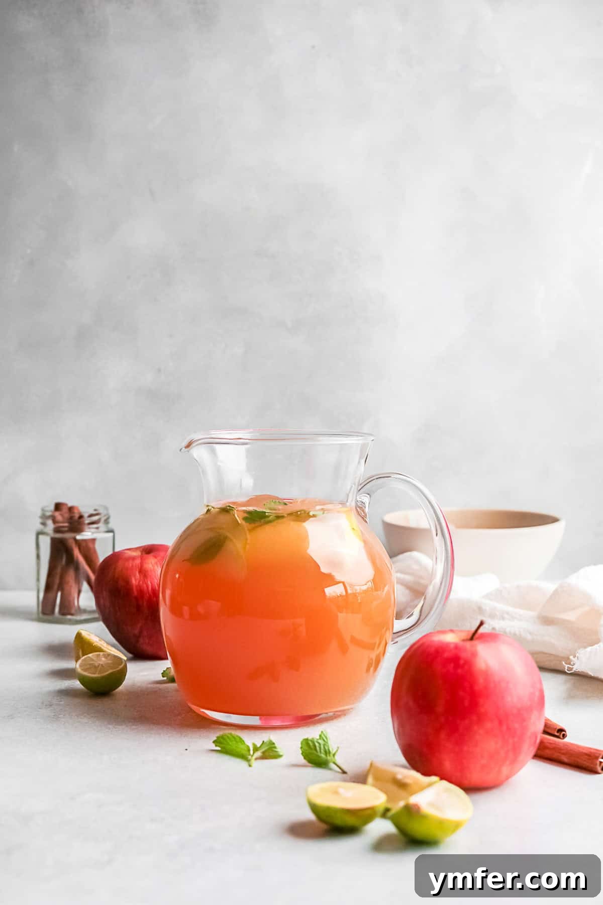 A large glass pitcher filled with pre-mixed Apple Cider Mojito, ready to be served, alongside a glass with fresh mint and lime.