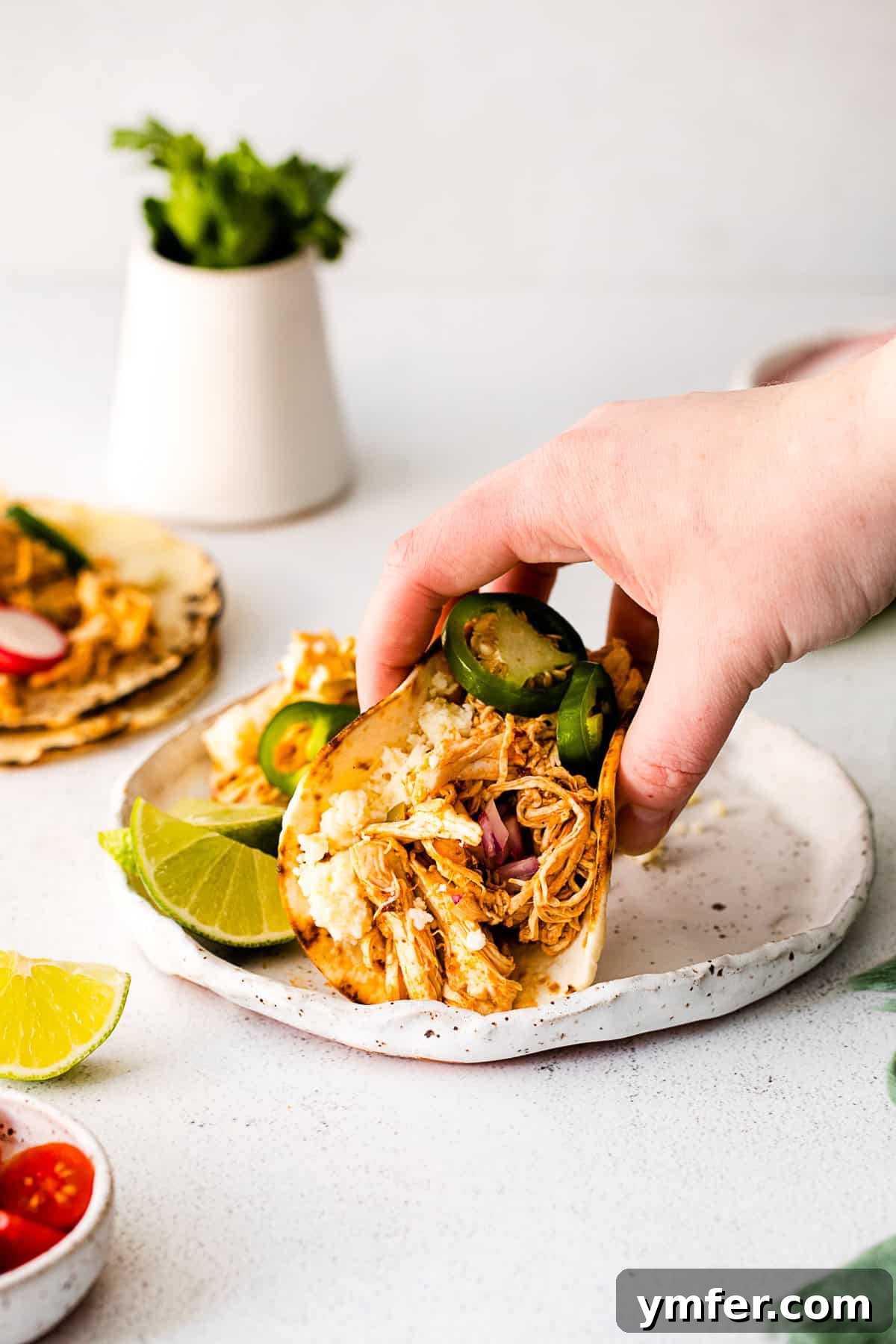Effortless Instant Pot Chicken Tacos 6 A hand reaching for an Instant Pot Chicken Taco, garnished with cilantro and a lime wedge, presented on a white plate.