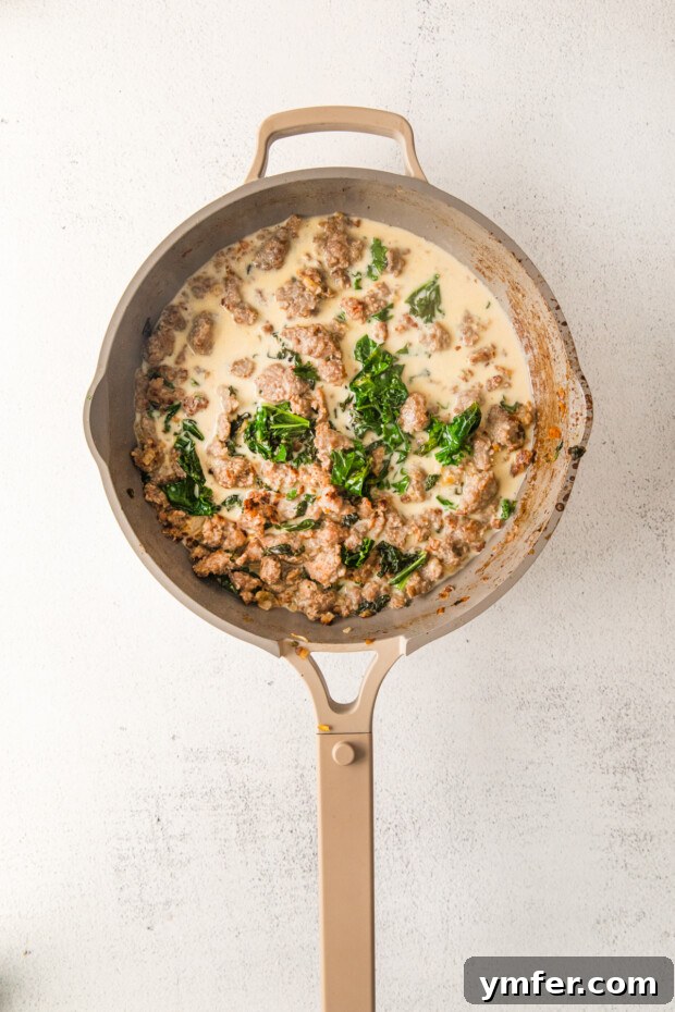 Autumn Butternut Squash Sausage Pasta 5 Heavy cream being poured into a skillet with browned sausage and wilted kale, forming the rich pasta sauce.