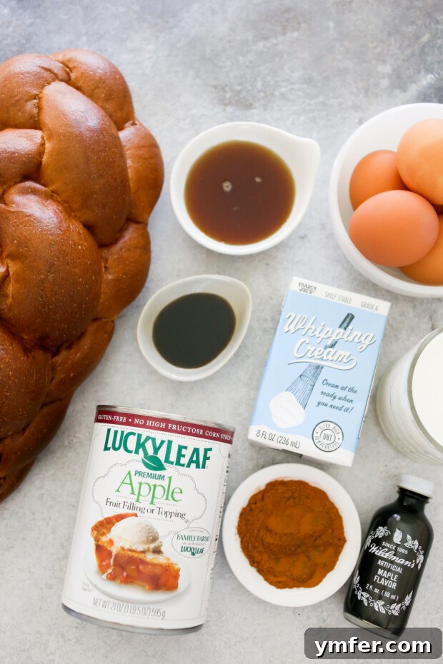 Ingredients for Apple Pie Baked French Toast including challah bread, maple syrup, eggs, vanilla extract, milk, heavy cream, and a can of apple pie filling, neatly arranged on a rustic wooden table.