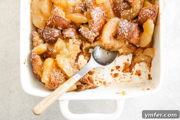 A serving of Apple Pie Baked French Toast in a white dish with a spoon, showing the rich apple filling and custardy bread.