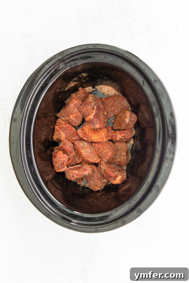 Melt-in-Your-Mouth Slow Cooker Beef Barbacoa 4 Uncooked Slow Cooker Beef Barbacoa ingredients, including spiced chuck roast cubes, sliced onions, and garlic, prepared and layered inside a slow cooker before cooking.