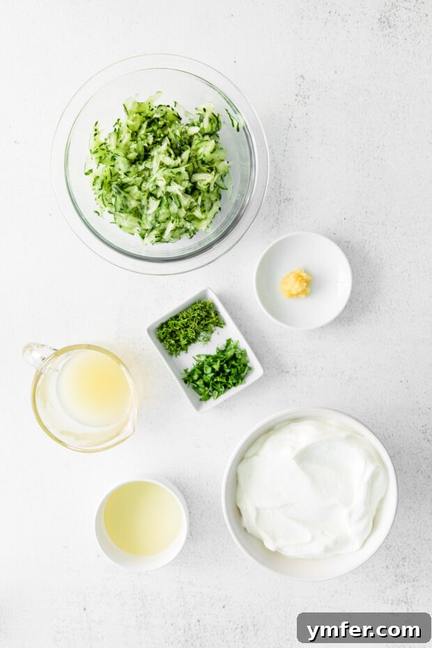 Refreshing Cucumber Yogurt Dip 3 Individual ingredients for Tzatziki sauce laid out on a table: shredded cucumber, a bowl of Greek yogurt, and a selection of fresh herbs.