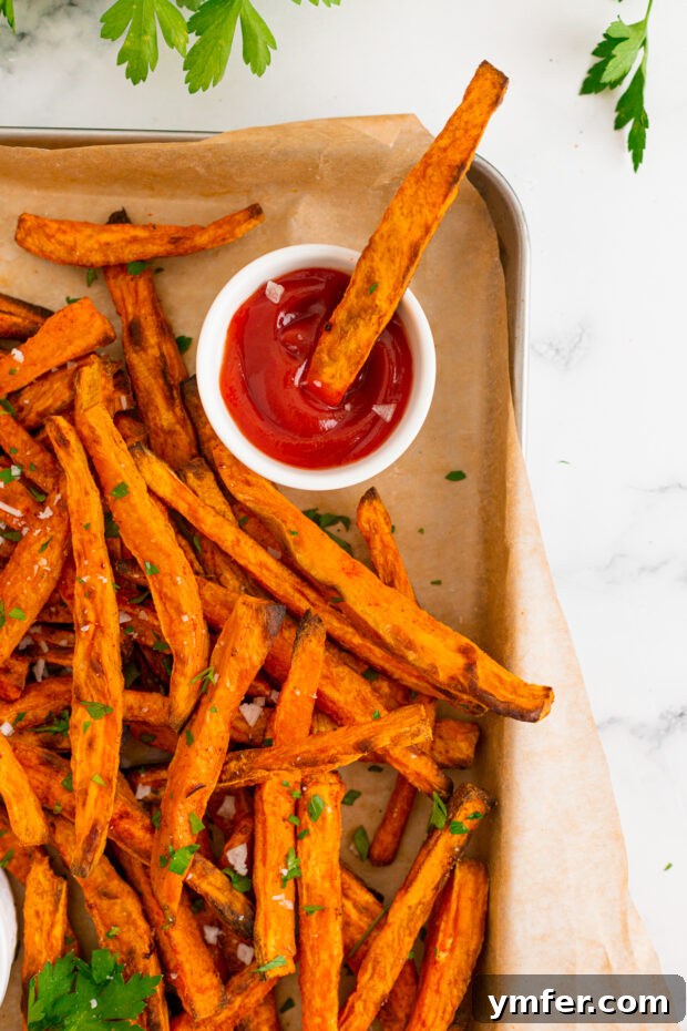 Gourmet Sweet Potato Fries 7 A single sweet potato fry being dipped into a dollop of bright red ketchup on a white plate, showcasing its golden color.