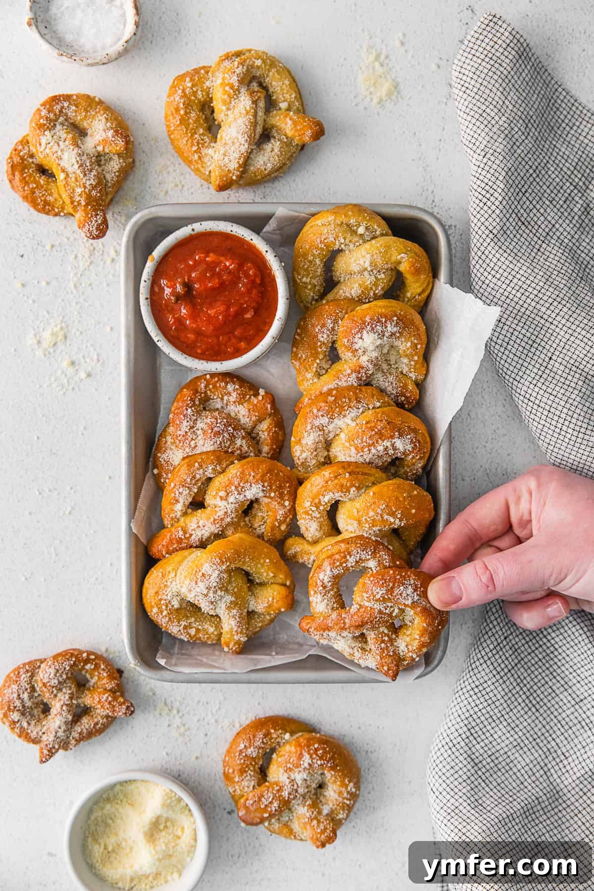 A hand reaching to pick up a warm garlic parmesan soft pretzel from a tray, emphasizing its fresh and inviting appeal.