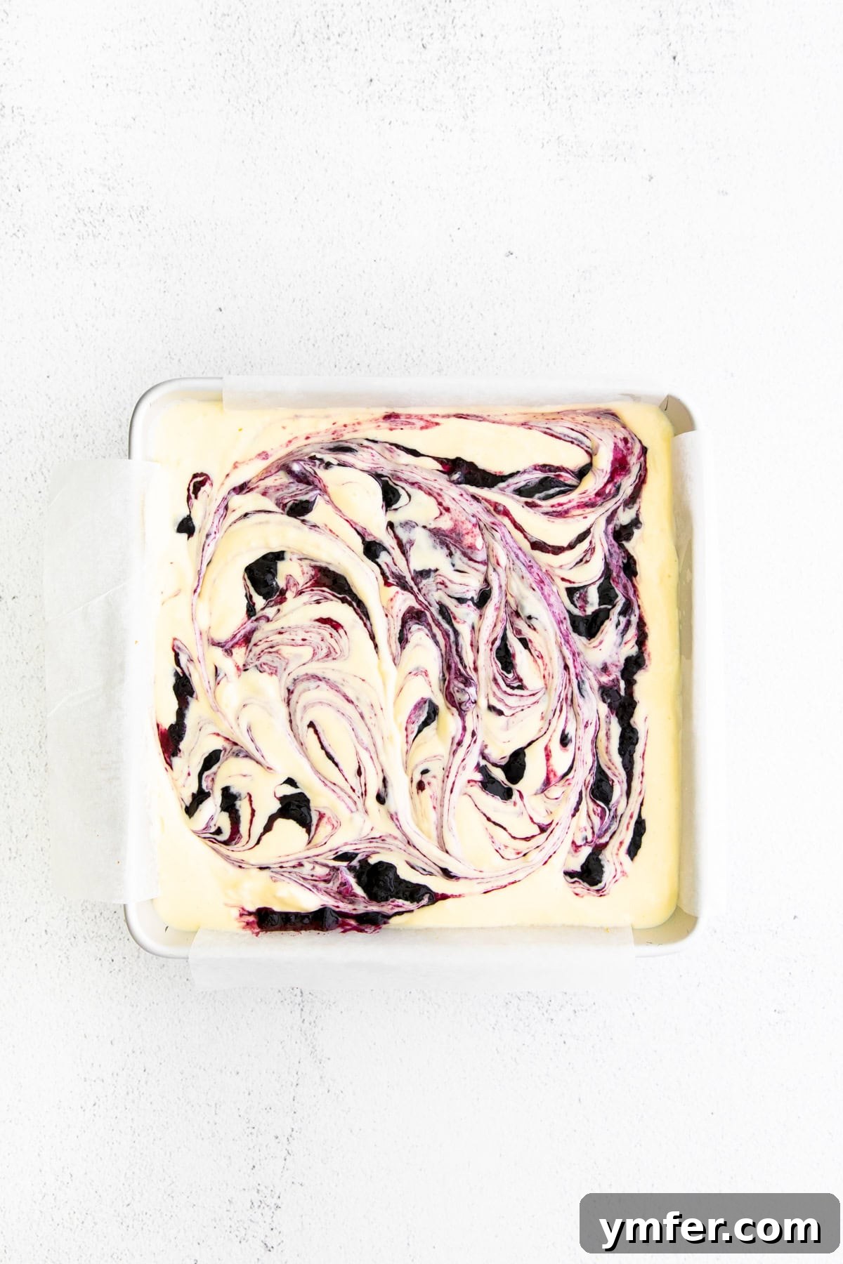 Luscious Blueberry Cheesecake Bars 5 Unbaked blueberry cheesecake bars in a baking pan, showing the swirled blueberry jam and scattered fresh blueberries on top.