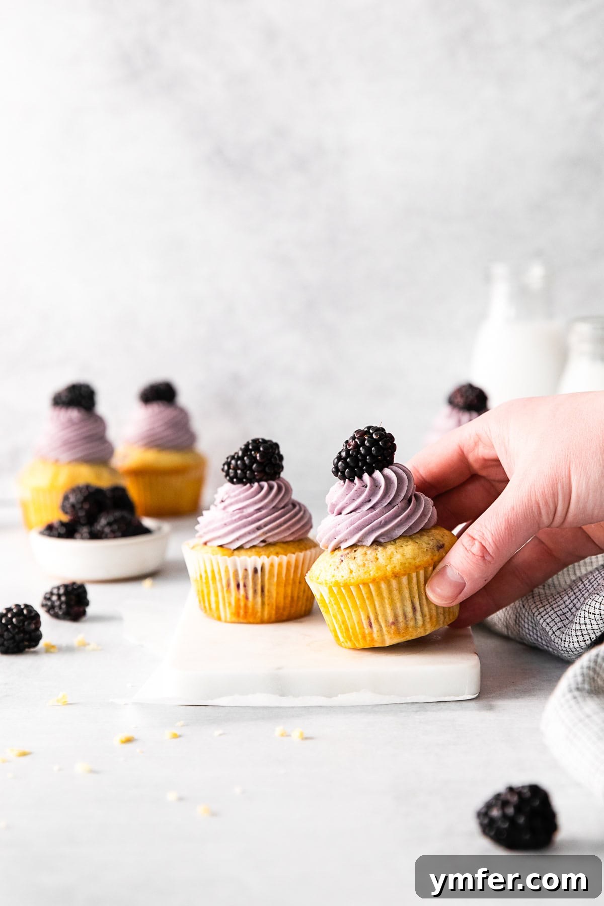 Blackberry Bramble Cupcakes 10 A hand reaching to pick up a frosted Blackberry Cupcake from a white serving tray.