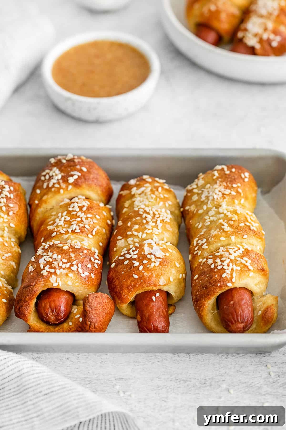 Golden Pretzel Franks 7 Two soft pretzel dogs topped with a sprinkle of sesame seeds and a generous amount of coarse salt, presented on a baking sheet.