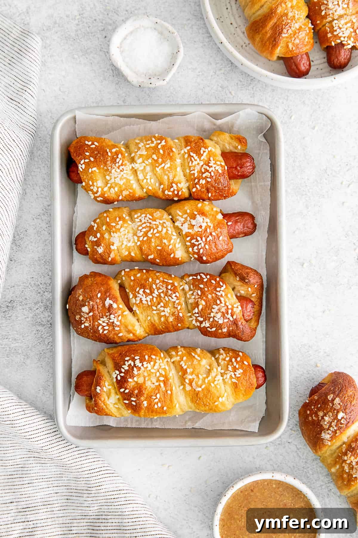 Golden Pretzel Franks 8 Freshly baked Soft Pretzel Dogs arranged on a sheet pan, with a small bowl of dipping sauce on the side.