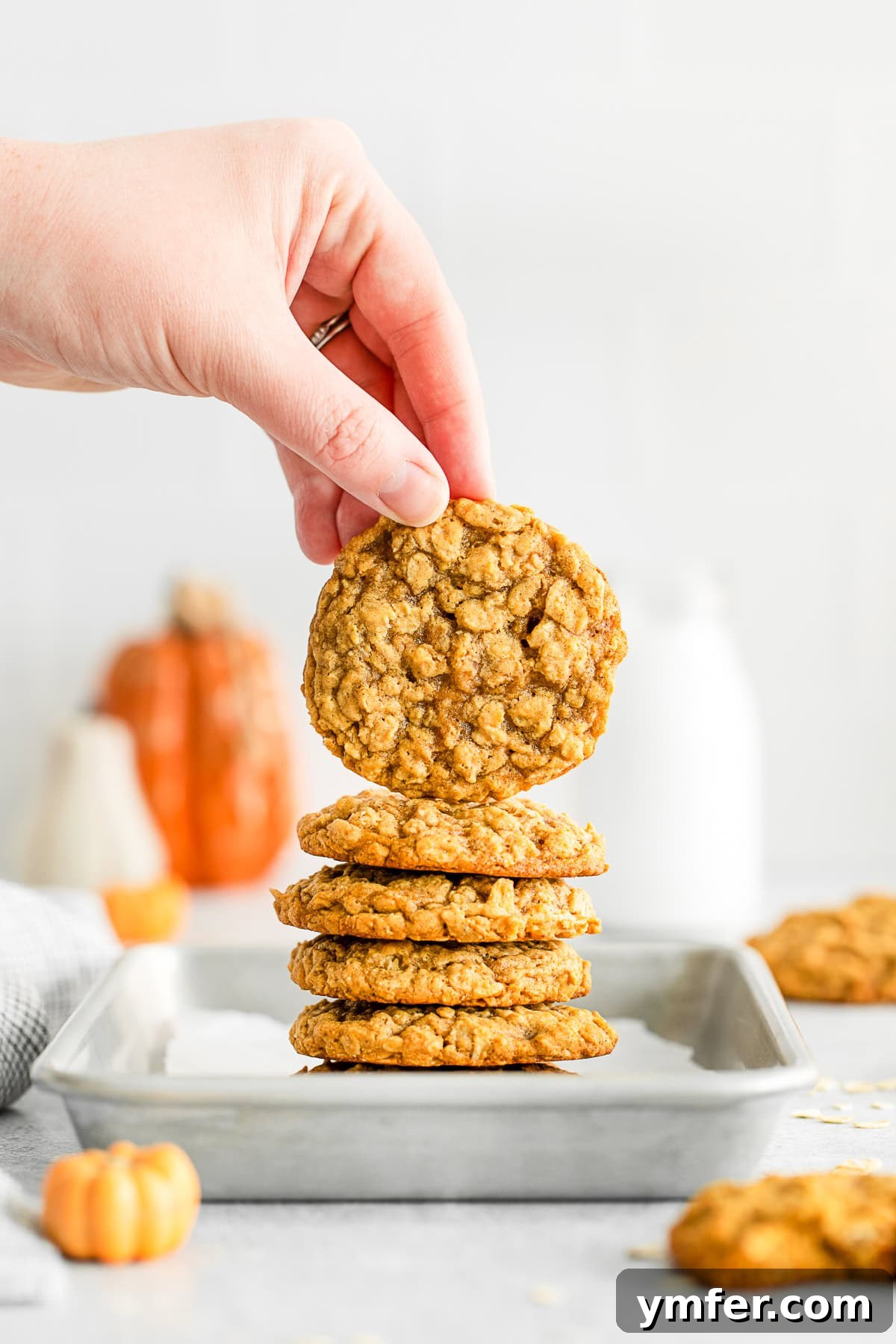 Autumn Spice Oatmeal Delights 8 Stacked Pumpkin Oatmeal Cookies on a sheet pan with a hand holding one of the cookies.