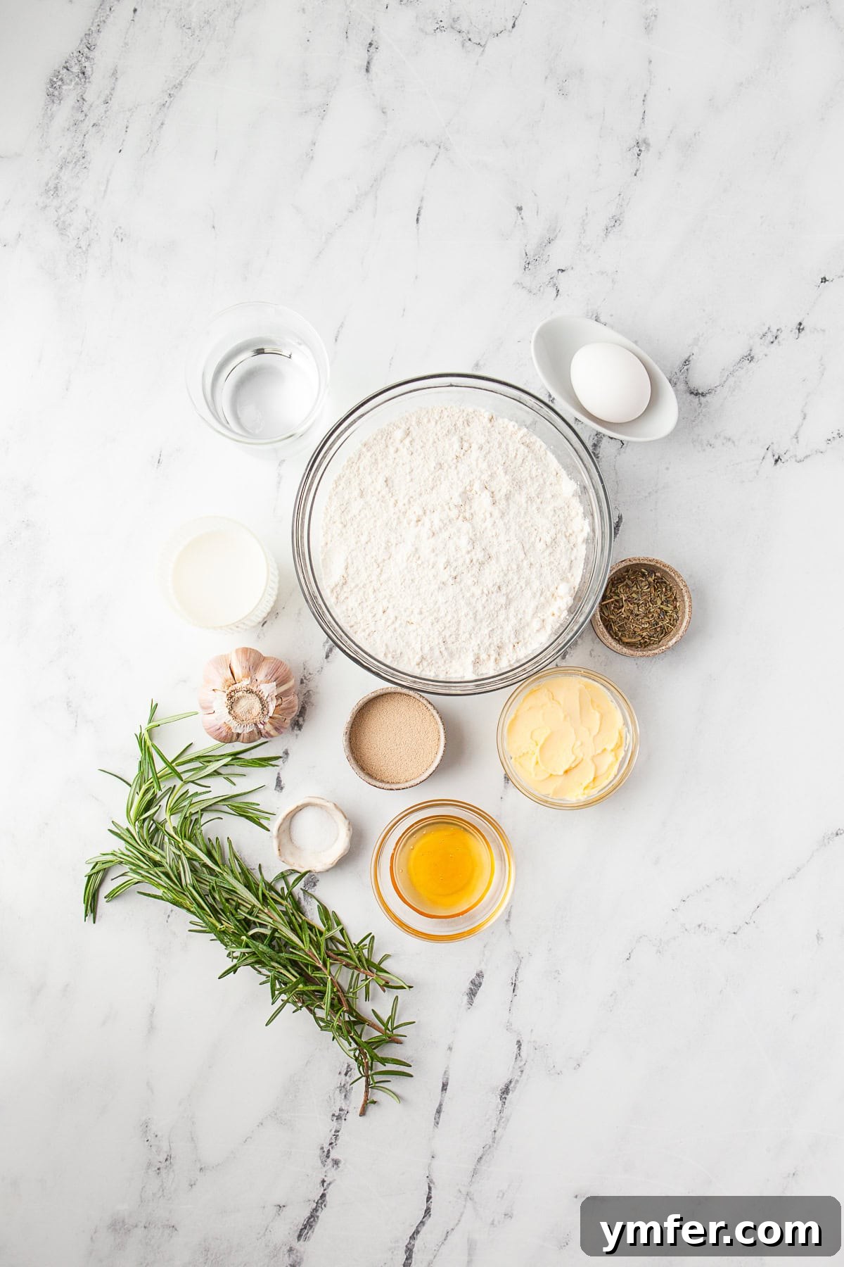 Warm Garlic Herb Rolls 3 A collection of fresh ingredients for garlic herb dinner rolls laid out on a kitchen counter: milk, butter, honey, yeast, eggs, garlic, rosemary, and flour.
