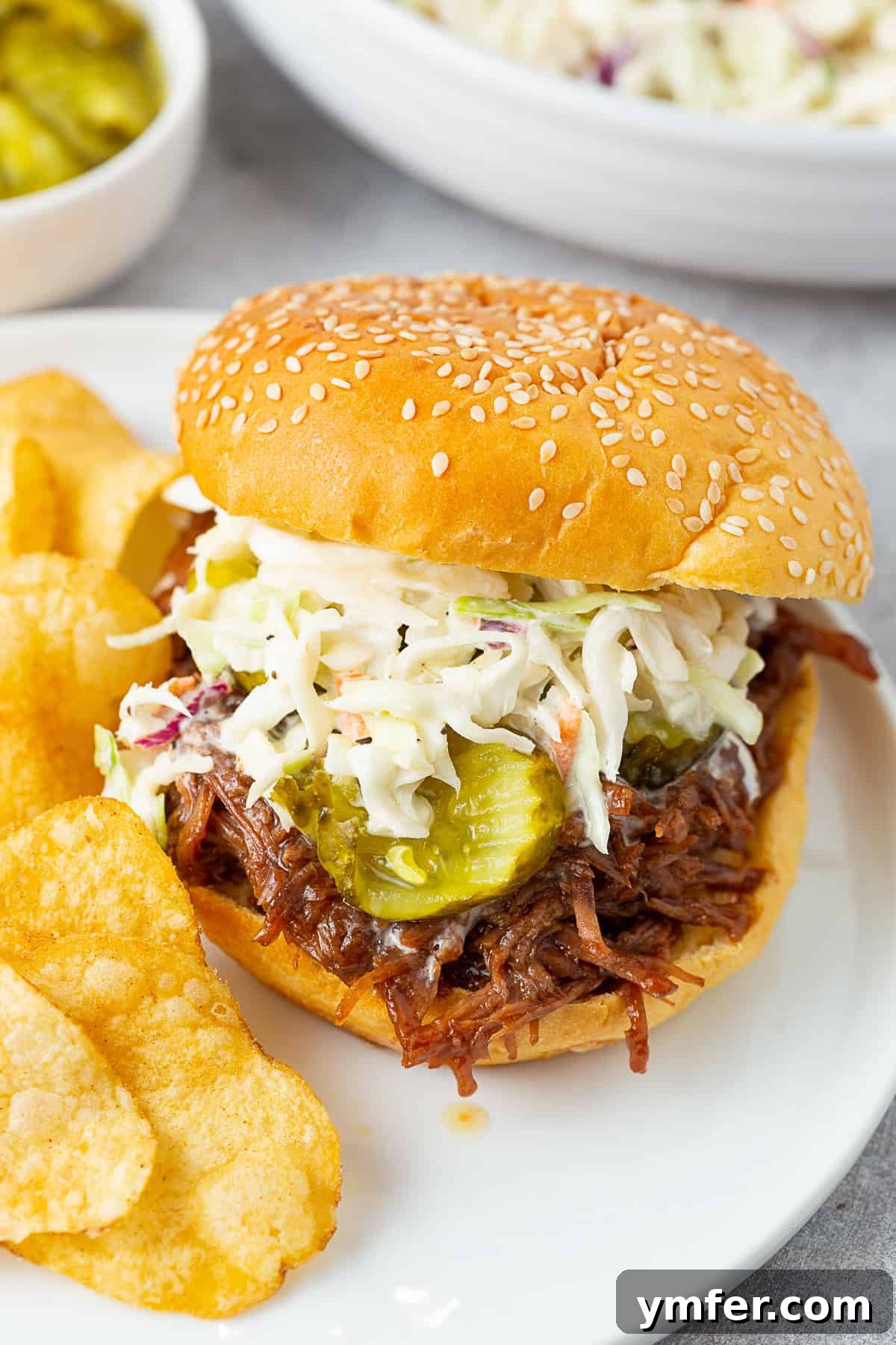 Smoky Pulled Beef Sandwiches 4 BBQ Shredded Beef (Sandwiches) topped with coleslaw and pickles, served on a plate.
