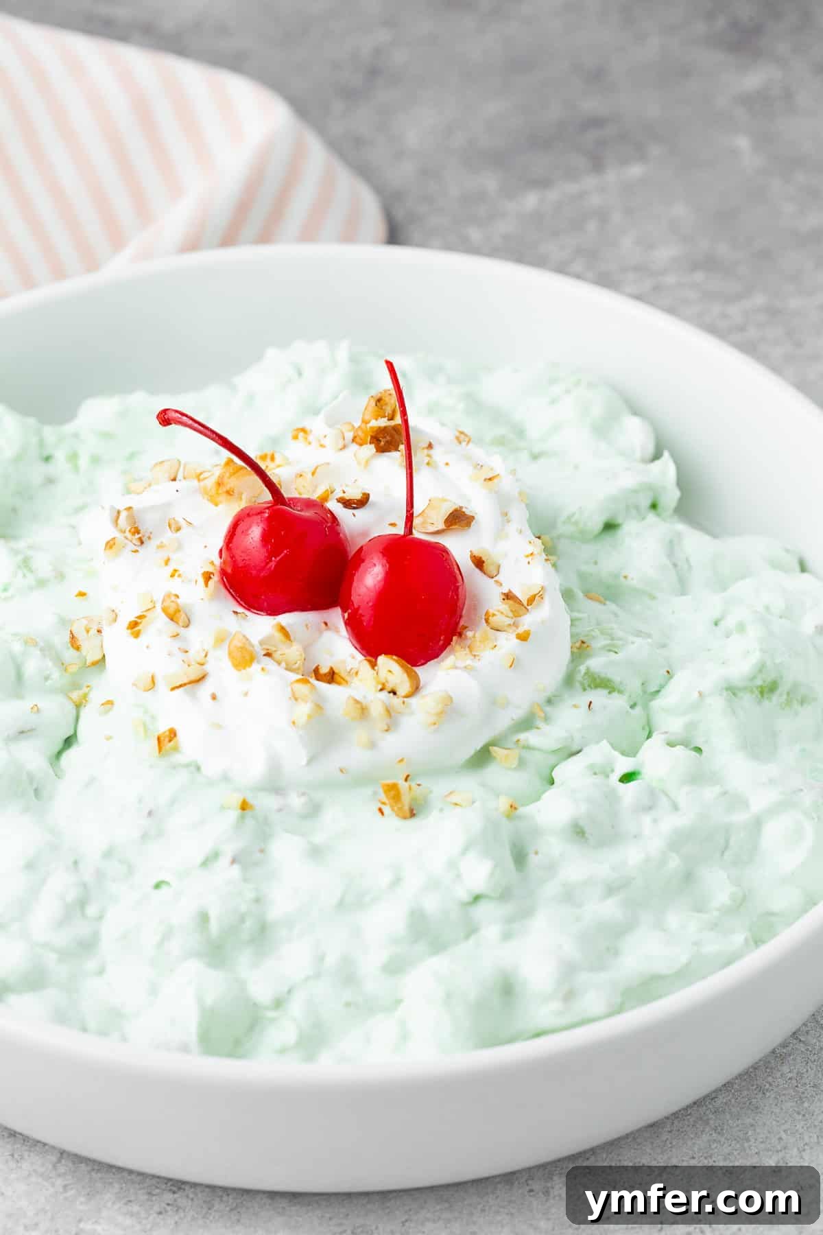 Emerald Pineapple Dream 2 Watergate Salad in a white bowl topped with cherries. A vibrant green, fluffy dessert ready to be enjoyed.