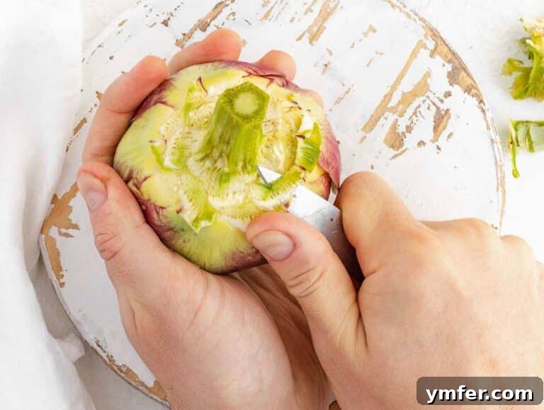 Hands meticulously cleaning the stem of an artichoke with a paring knife for a smoother texture.