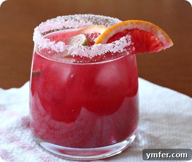 Vibrant Blood Orange Margarita cocktail in a salt-rimmed glass, garnished with fresh blood orange and lime slices, against a light background.