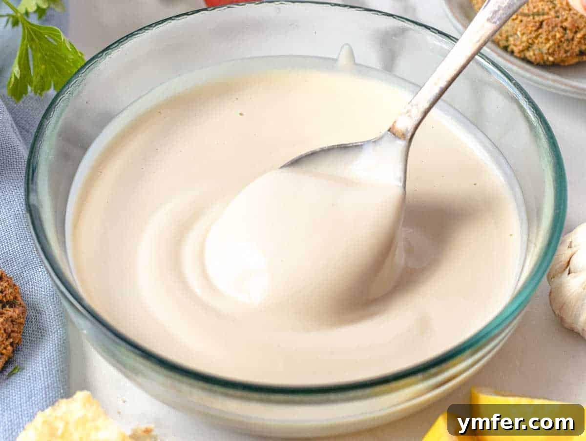 Silky Tahini Drizzle 4 A close-up of creamy tahini sauce in a clear glass bowl, with a small silver spoon resting inside.