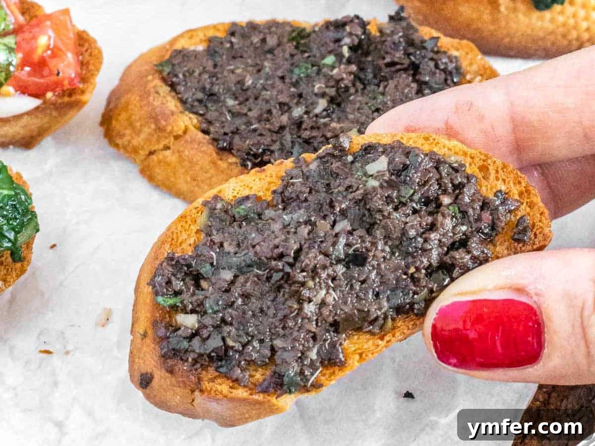 Rich Olive Spread 10 Hand reaching for a crostini with olive tapenade on a white plate