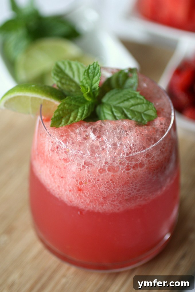 Strawberry Watermelon Agua Fresca by Nutmeg Nanny A close-up of a glass of strawberry watermelon agua fresca with ice, fresh mint, and lime wedges, set against a blurred background, highlighting its inviting and vibrant qualities.