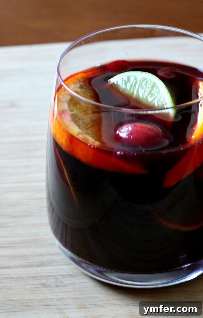 A hand pouring red wine sangria from a pitcher into a glass filled with ice and fresh fruit, surrounded by more fruit on a wooden surface.