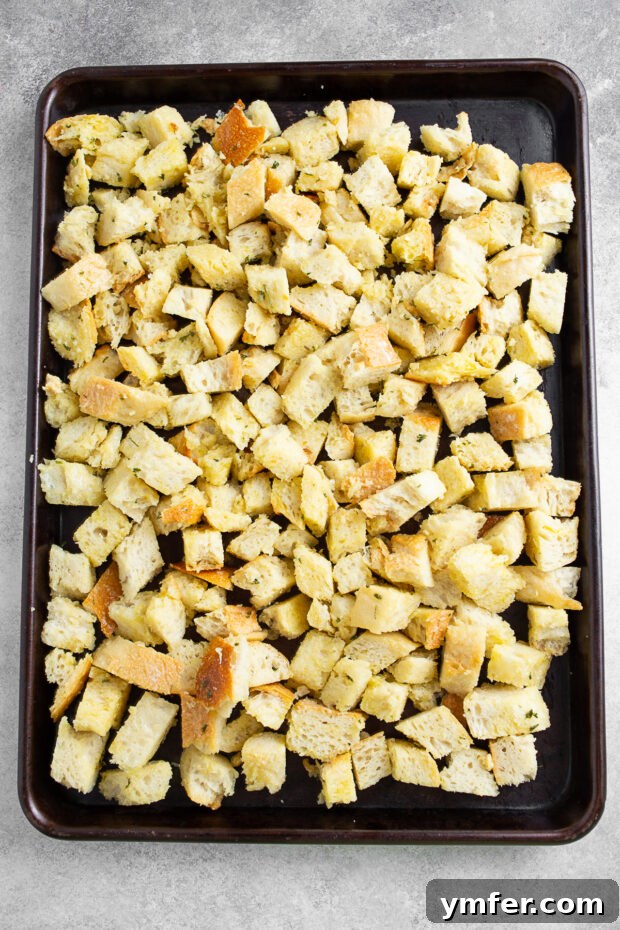 Seasoned croutons awaiting their transformation in the oven. Uncooked homemade croutons, thoroughly coated in garlic butter and herbs, spread out on a baking sheet before going into the oven.