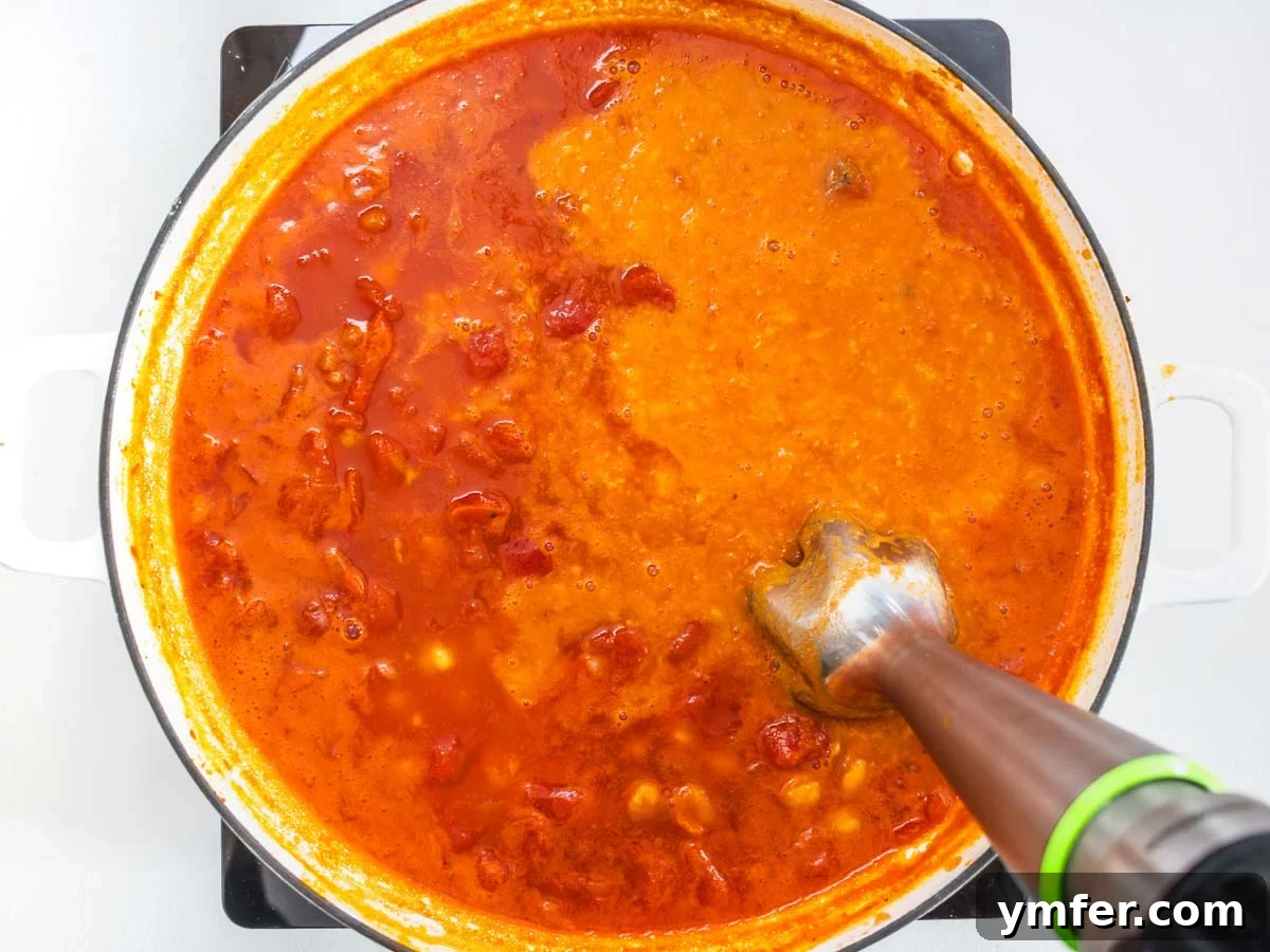 Hearty Chickpea Pasta 7 An immersion blender actively blending chickpeas and tomato sauce in a skillet, creating a creamy texture.