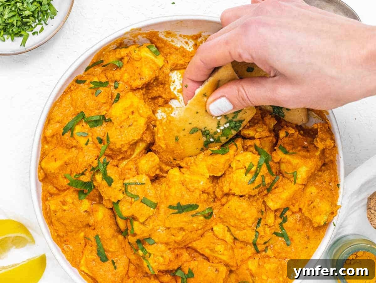 Velvety Vegan Butter Chicken 5 A hand holding a piece of naan bread scooping creamy vegan butter chicken from a bowl, garnished with fresh herbs.