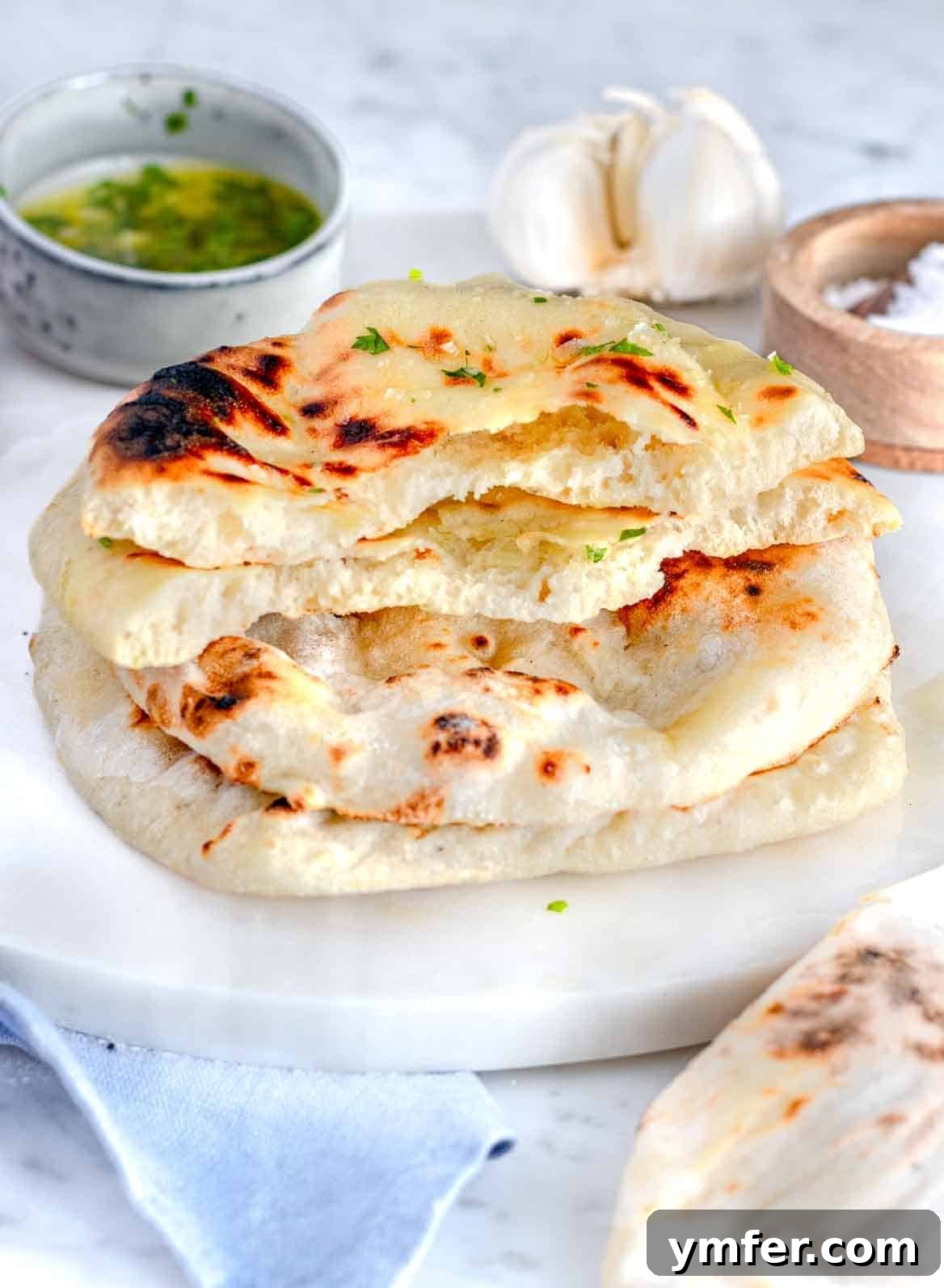 Pillowy Naan 2 Stacked homemade naan bread, ripped in half, on a rustic cutting board showcasing its fluffy interior and charred crust.