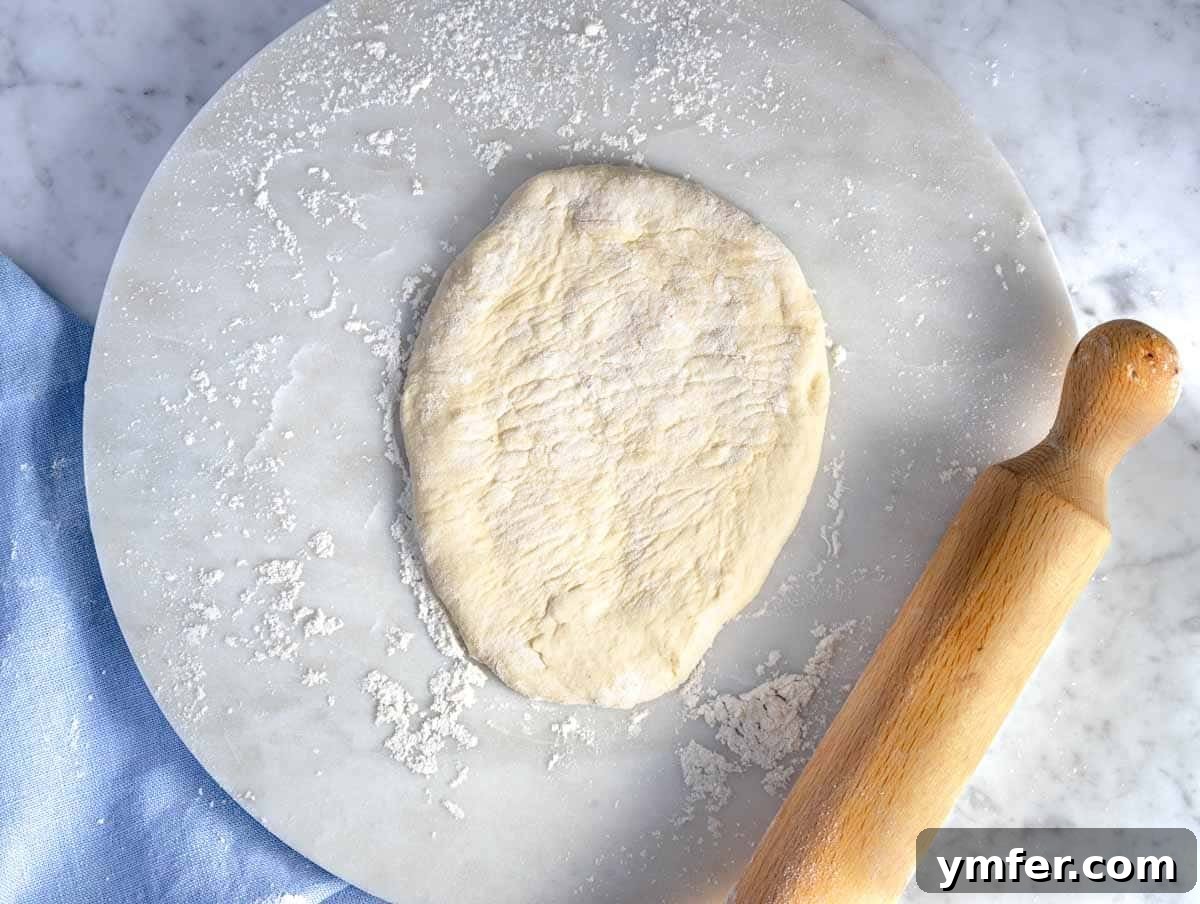 Pillowy Naan 11 Hands using a rolling pin to flatten a naan dough ball into an oval shape on a lightly floured surface.