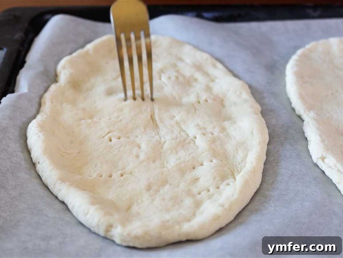 Pillowy Naan 14 Naan breads arranged on a baking sheet lined with parchment paper, pierced with a fork, ready for oven baking.