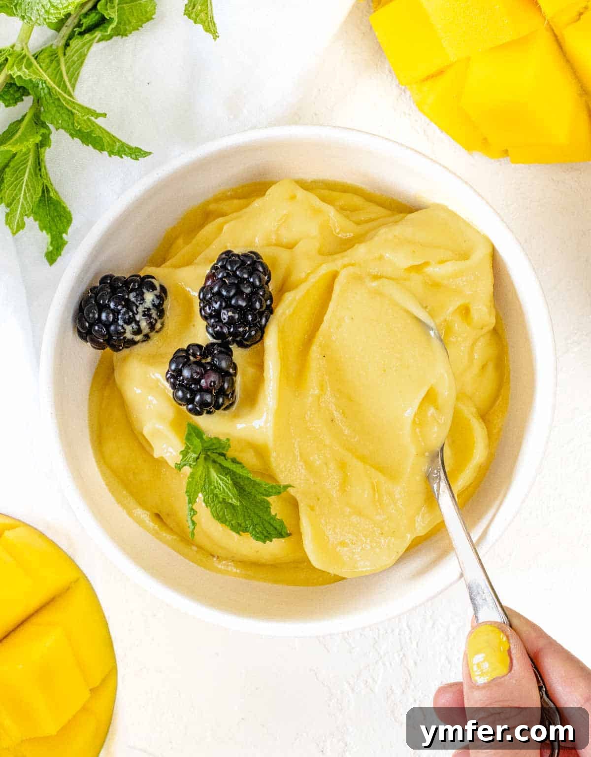 A hand holding a spoon scooping creamy mango sorbet from a bowl, garnished with fresh berries.