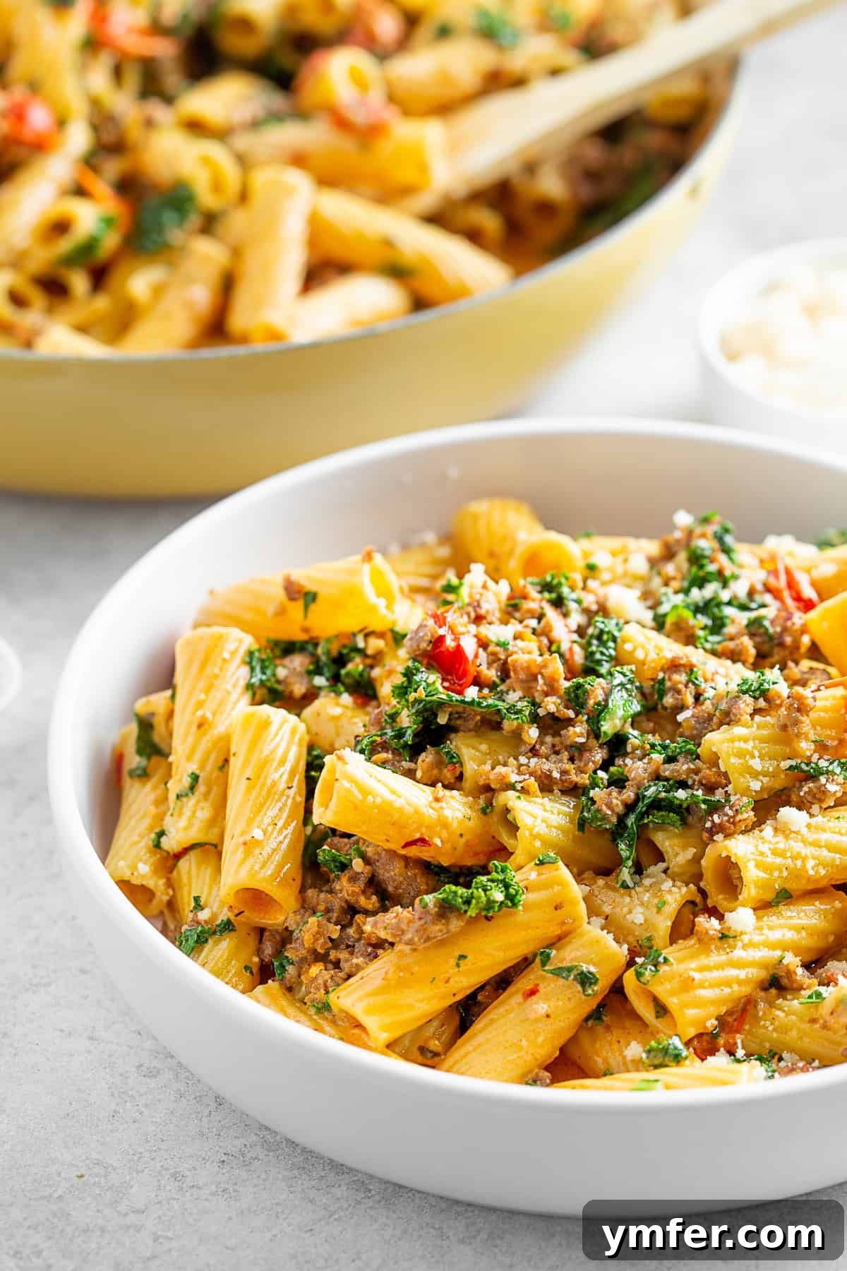 Italian Sausage Kale Pasta in a white bowl with a skillet of pasta in the back. 